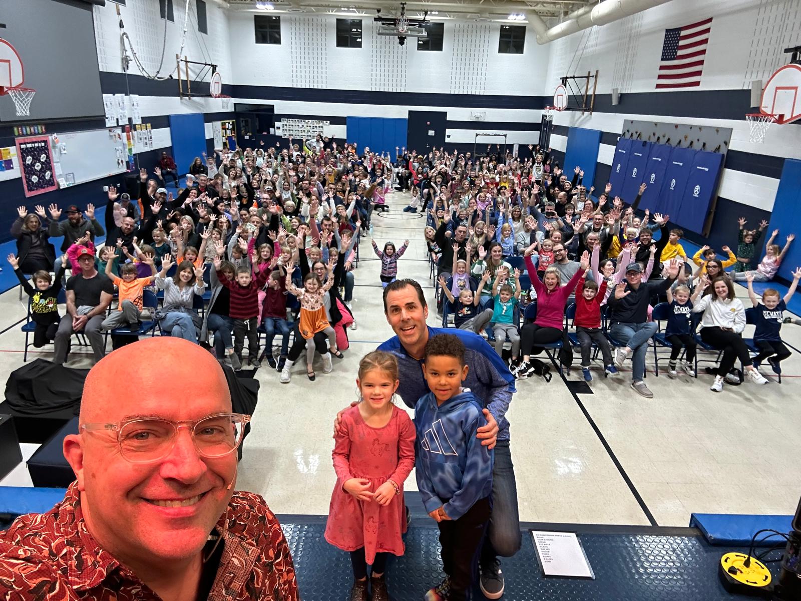 Tim on stage with the principal and two students, packed gym behind them with hands raised